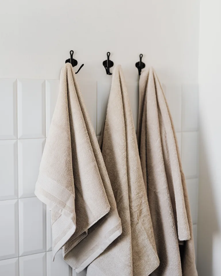 Three beige towels hanging on individual hooks on a tiled bathroom wall