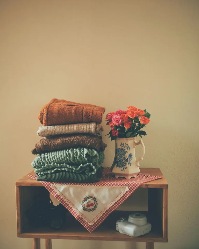 Stack of folded knit sweaters with a floral vase on a wooden stand