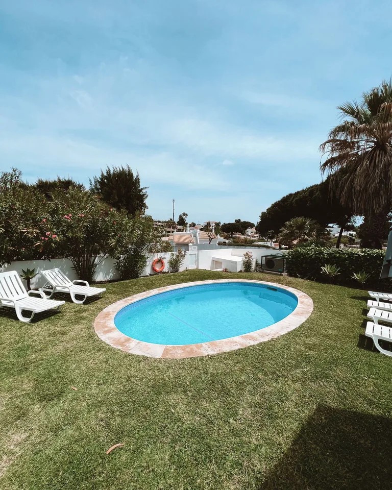 Oval swimming pool surrounded by lush greenery and lounge chairs under a blue sky